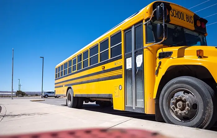 A yellow school bus parked on a paved lot under a clear blue sky, with a distant car and landscape visible in the background.
